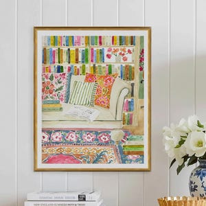 May include: A framed watercolour print shows a reading nook with a sofa, cushions, and a bookshelf. The print is in a gold frame and is displayed on a wooden dresser. A stack of books, a gold bowl, and a blue and white vase with flowers are also on the dresser.