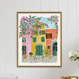 May include: A framed watercolor painting depicting a Mediterranean street scene with colorful yellow, pink, and orange buildings, green shutters, a cobblestone ground, and vibrant pink bougainvillea flowers cascading over the architecture.
