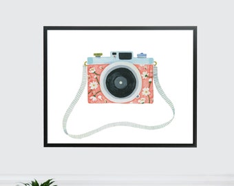 Könnte beinhalten: Ein gerahmter Druck einer Aquarellillustration einer rosa und blauen Kamera mit einem Blumenmuster. Die Kamera hat einen Riemen und hängt an einer weißen Wand.