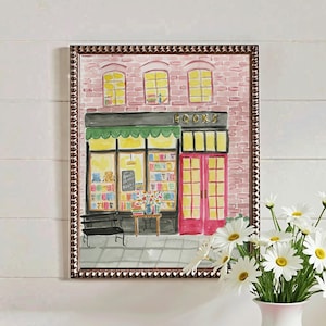 May include: Watercolor painting of a bookstore with a green awning, red door, and the word "BOOKS" above the entrance. The painting is in a decorative frame. A white vase with daisies sits on a table with a pink book.
