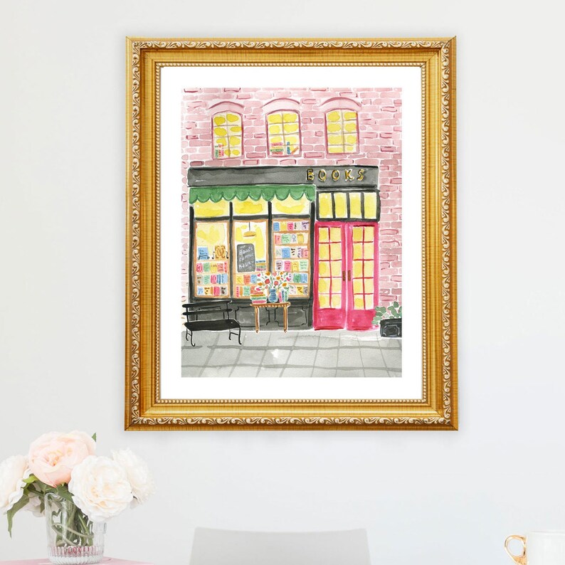 May include: Watercolor painting of a bookstore facade with a gold frame. The building has a brick exterior, green awning, and red doors. The word "BOOKS" is above the window. A black bench and table with flowers are in front.