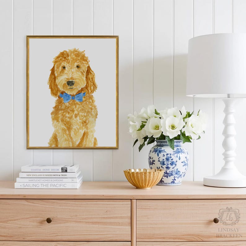 May include: A framed wall art print featuring a watercolor-style illustration of a golden-brown doodle dog with a bright blue bow tie. The dog is depicted from the chest up against a light gray background, enclosed in a thin gold-colored frame.