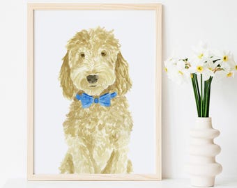 May include: Watercolor painting of a golden doodle dog wearing a blue bow tie, framed in a light wood frame. The dog is centered against a white background. A vase of white flowers sits to the right of the framed artwork.