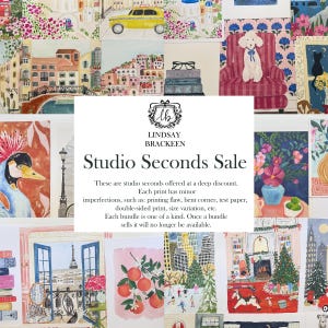 May include: A collection of colorful watercolor art prints featuring various scenes including cityscapes, floral arrangements, animals, and holiday themes. A central white sign displays the text LINDSAY BRACKEEN Studio Seconds Sale.