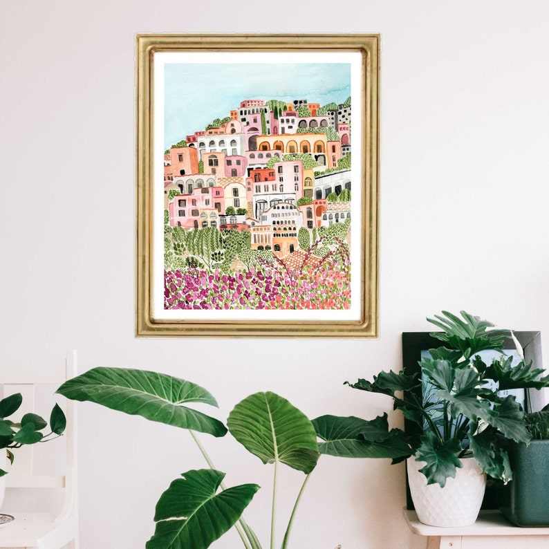May include: A watercolor painting of a colorful Italian village with pink, white, and yellow buildings. The buildings are clustered together on a hillside with green trees and bushes. The painting is framed in a gold frame and is hanging on a white wall.