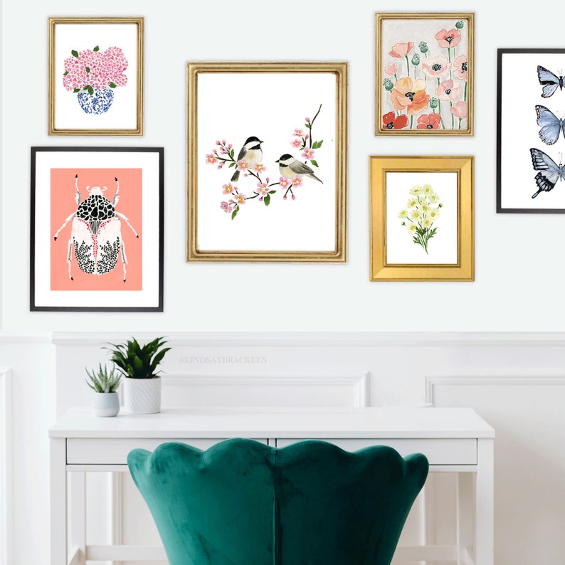 May include: A gallery wall of framed art prints, including floral and insect designs. The prints showcase a pink and blue floral arrangement, a beetle, two birds on a branch, and poppy flowers. The frames are gold and black, displayed above a white desk.