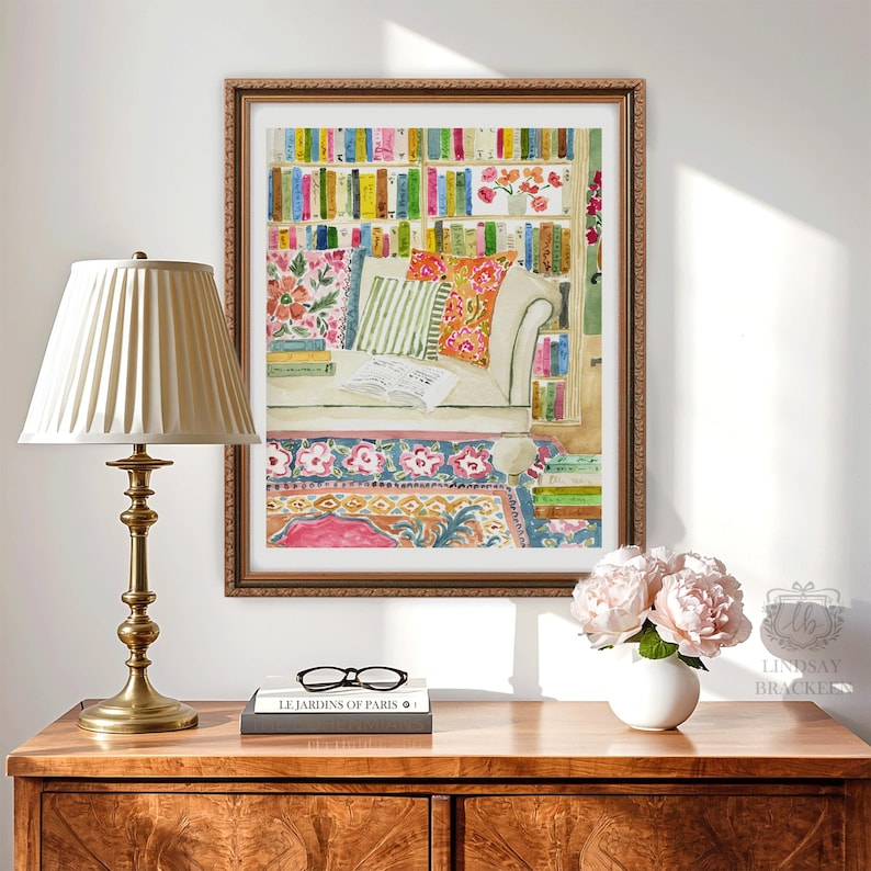 May include: A framed watercolour print shows a reading nook with a sofa, cushions, and a bookshelf with colourful books. A brass lamp, books, and a vase of flowers are on a wooden table in front of the print. The print is by Lindsay Brackers.