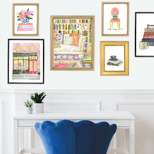May include: Watercolor art prints in gold frames, featuring stacks of books, a bookstore, and floral arrangements. The prints are displayed on a white wall above a white desk and a blue velvet chair.