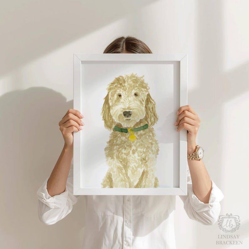 May include: A framed watercolor painting of a light brown, shaggy-haired dog with a green collar and a small yellow tag. The dog is depicted in a seated position against a plain white background, captured within a simple white rectangular frame.