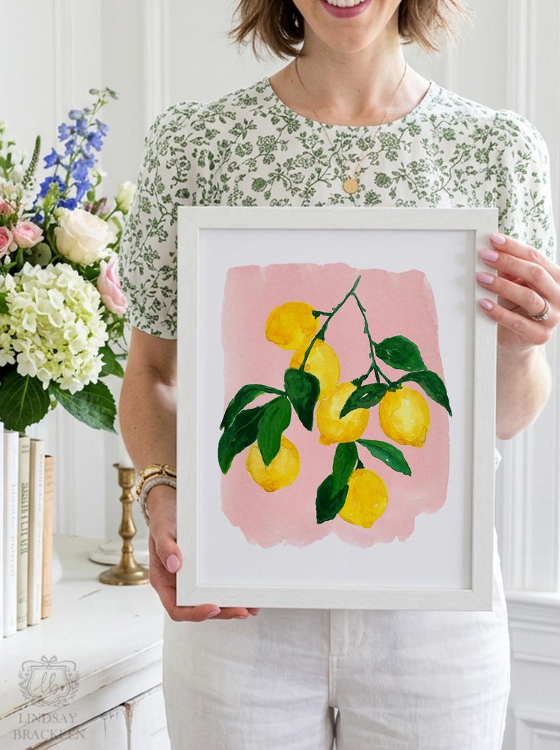 Op de afbeelding: Een ingelijste kunstdruk met een illustratie van gele citroenen aan groene, bebladerde takken tegen een zachtroze achtergrond. Het kunstwerk zit in een eenvoudige witte lijst met een strak, rechthoekig ontwerp.