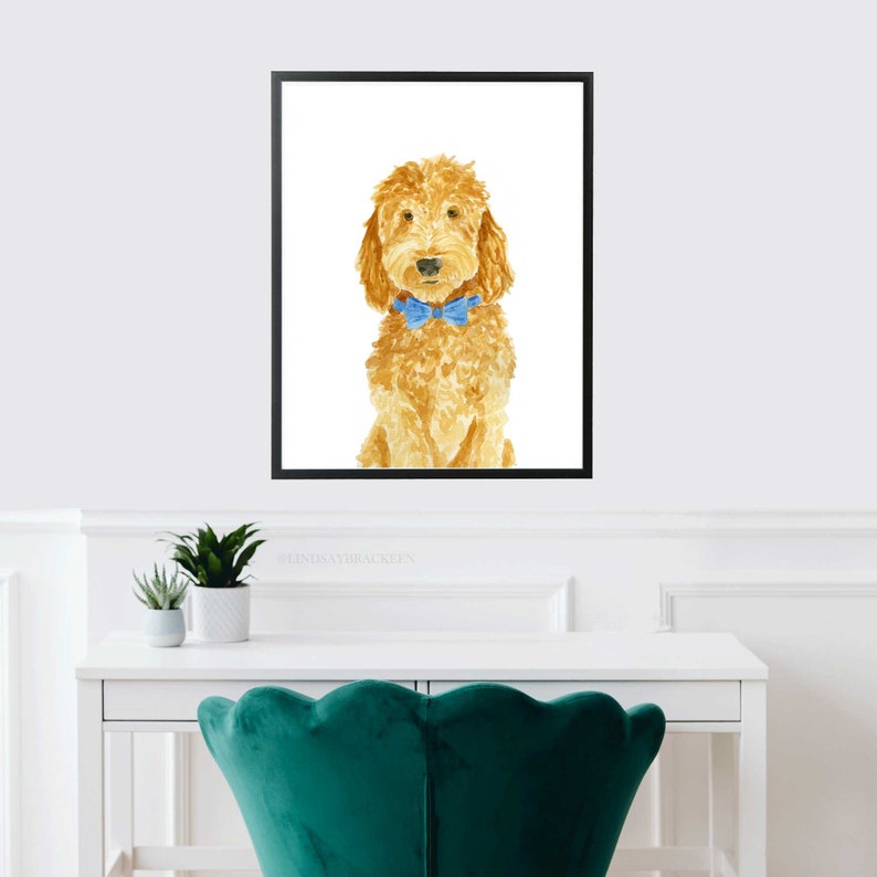 May include: A framed watercolor painting of a golden-colored curly-haired dog wearing a bright blue bow tie. The dog is depicted in a seated position against a plain white background, enclosed within a simple black rectangular frame.