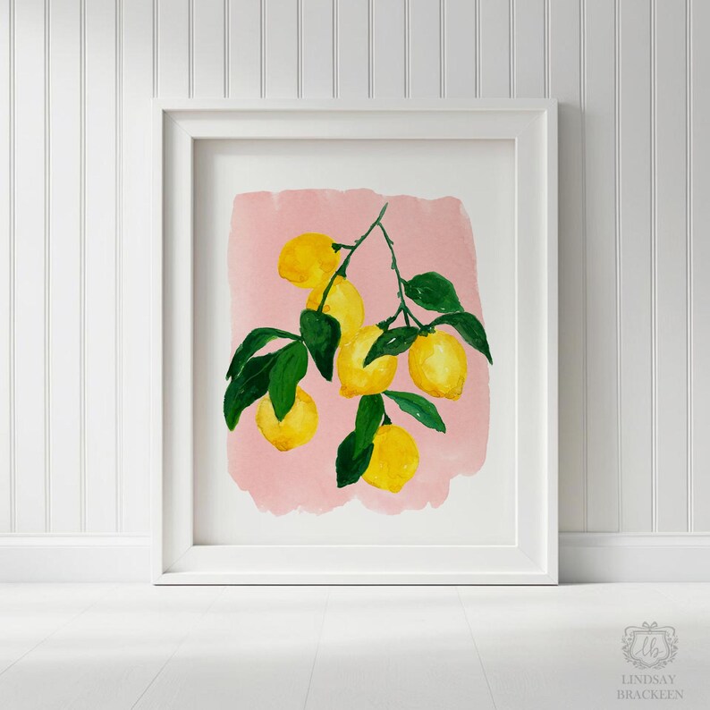 Op de afbeelding: Een ingelijste kunstdruk met een aquarelillustratie van een citroentak met vijf felgele citroenen en donkergroene bladeren, gecentreerd op een zachtroze rechthoekige achtergrond in een strakke witte lijst.