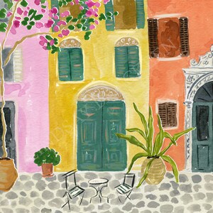May include: A watercolor painting of a cobblestone street scene featuring colorful buildings in pink, yellow, and orange. The scene includes green shutters, arched doorways, a potted plant, a small table with two chairs, and a flowering tree.
