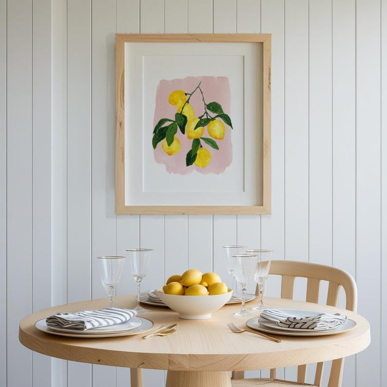 May include: A dining room scene featuring a round wooden table set for a meal. A framed watercolor painting of lemons hangs on the white wall. The table is set with plates, napkins, glasses, and a bowl of lemons.