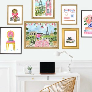 May include: A white desk with a gold metal chair, a laptop, a lamp, and a potted succulent plant. There are six framed watercolor paintings on the wall above the desk. The paintings feature a woman with a bow in her hair, a Parisian cafe scene, a cup of tea, a stack of books, and a pair of glasses. The text '@lindsaybracken' is written on the wall below the paintings.