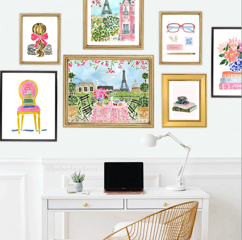 May include: A white desk with a gold metal chair, a laptop, a lamp, and a potted succulent plant. There are six framed watercolor paintings on the wall above the desk. The paintings feature a woman with a bow in her hair, a Parisian cafe scene, a cup of tea, a stack of books, and a pair of glasses. The text '@lindsaybracken' is written on the wall below the paintings.