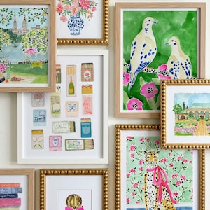 Op de afbeelding: Een galerijwand met meerdere ingelijste aquareldrukken. De collectie bevat illustraties van een bloemenboeket, een parksc&egrave;ne, luciferdoosjes, twee vogels op een tak, een luipaard met een roze strik, boeken en een persoon met een strik.