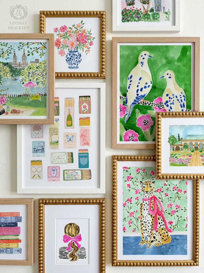 Op de afbeelding: Een galerijwand met meerdere ingelijste aquareldrukken. De collectie bevat illustraties van een bloemenboeket, een parksc&egrave;ne, luciferdoosjes, twee vogels op een tak, een luipaard met een roze strik, boeken en een persoon met een strik.