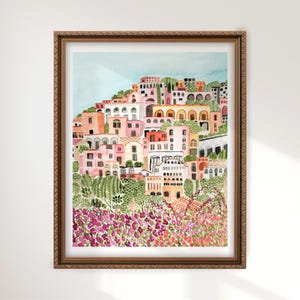 May include: Watercolor painting of a coastal Italian village in a brown wooden frame. The artwork depicts colorful buildings on a hillside, with pink, orange, and white facades. Lush green foliage and vibrant purple flowers are in the foreground.