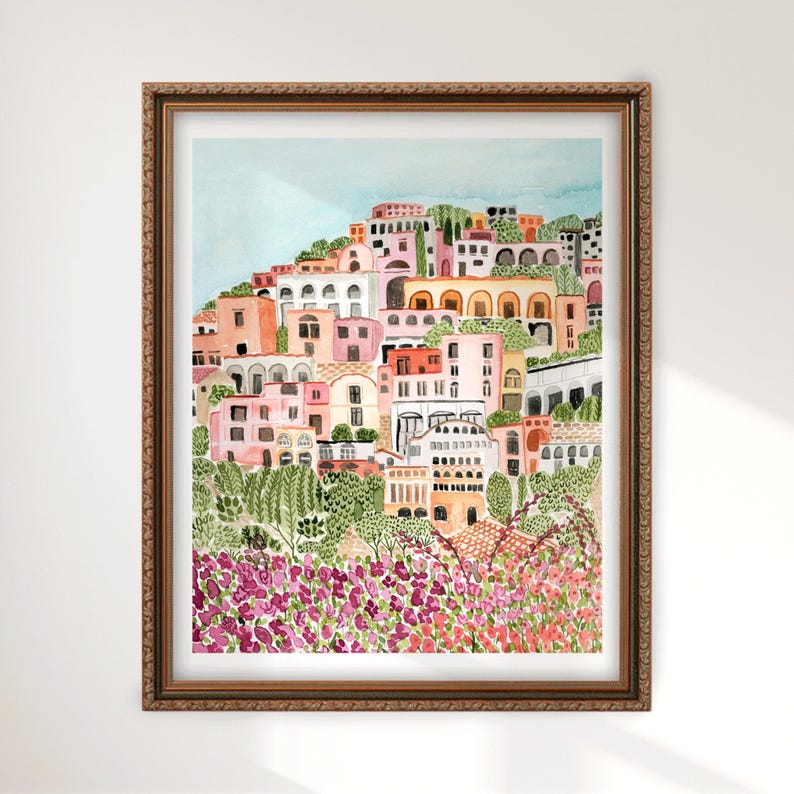 May include: Watercolor painting of a coastal Italian village in a brown wooden frame. The artwork depicts colorful buildings on a hillside, with pink, orange, and white facades. Lush green foliage and vibrant purple flowers are in the foreground.