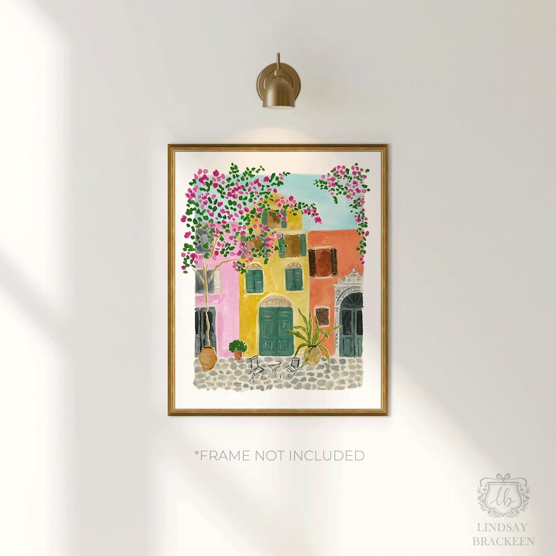 May include: A watercolor print featuring a street scene with pink, yellow, and orange buildings, a cobblestone ground, and vibrant pink bougainvillea flowers. The artwork is displayed in a gold frame with the text *FRAME NOT INCLUDED below it.