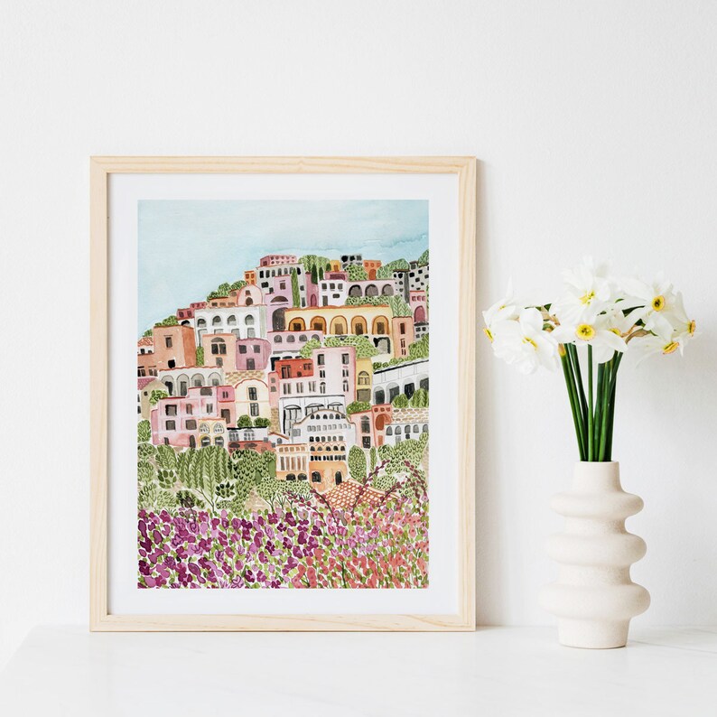 May include: A watercolor painting of a colorful Italian village with a hillside of purple flowers in the foreground. The village is painted in shades of pink, yellow, orange, and green. The painting is framed in a light wood frame.
