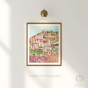May include: A framed watercolor print of a coastal town on a hillside, featuring buildings in shades of pink, peach, and white. The artwork is set against a light blue sky and framed in a gold-colored frame. The text "*FRAME NOT INCLUDED" is visible.