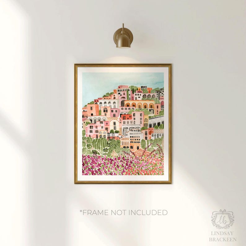 May include: A framed watercolor print of a coastal town on a hillside, featuring buildings in shades of pink, peach, and white. The artwork is set against a light blue sky and framed in a gold-colored frame. The text "*FRAME NOT INCLUDED" is visible.