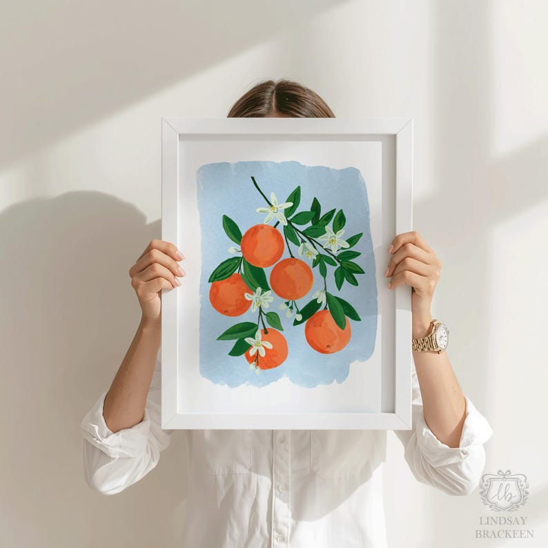 K&ouml;nnte beinhalten: Ein gerahmter Kunstdruck mit einer Aquarellillustration von Orangen an einem Zweig. Das Kunstwerk zeigt leuchtend orange Fr&uuml;chte, gr&uuml;ne Bl&auml;tter und zarte wei&szlig;e Bl&uuml;ten vor einem hellblauen Hintergrund. Der Druck wird von einer Person in einem wei&szlig;en Hemd gehalten.