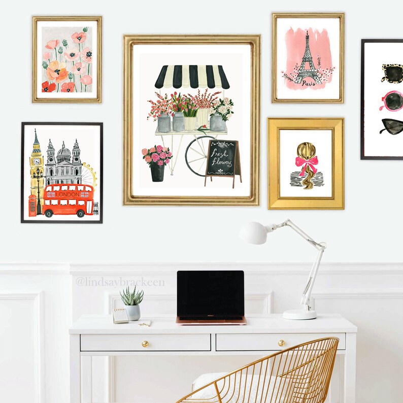May include: A white desk with a laptop, a lamp, and a potted plant. Above the desk are nine framed prints with various images, including a flower cart, the Eiffel Tower, and a red double-decker bus. The text "@lindsaybracken" is written on the wall.