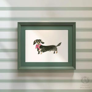 May include: A framed watercolor print of a dachshund dog wearing a pink scarf. The dog is black and tan with floppy ears and a curled tail. The frame is a muted green color, and the background is a striped wallpaper.