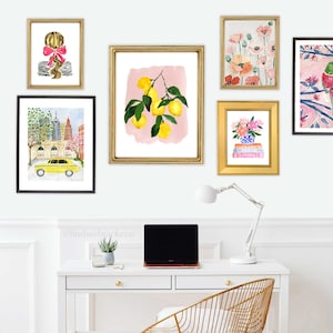 May include: A collection of framed watercolor art prints. The prints feature a variety of subjects, including a lemon branch, flowers, a New York City scene with a yellow taxi, and a portrait of a girl with a pink bow. The frames are gold and black.