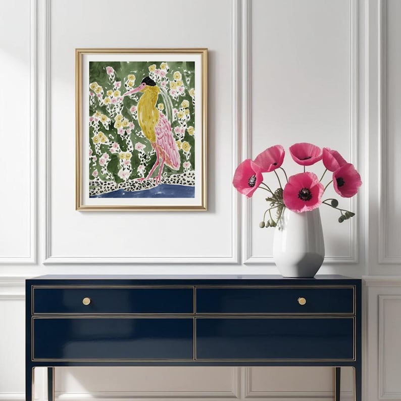 May include: A framed watercolor print featuring a bird with yellow and pink plumage, set against a green floral background. The artwork is displayed above a navy blue dresser with gold accents, complemented by a vase of pink flowers.