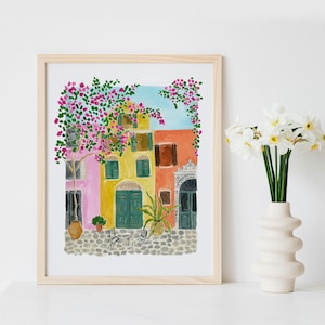 May include: A framed watercolor print depicting a Mediterranean street scene with pink, yellow, and orange buildings, green shutters, a cobblestone ground, a small table with two chairs, potted plants, and a large tree with pink flowers against a blue sky.