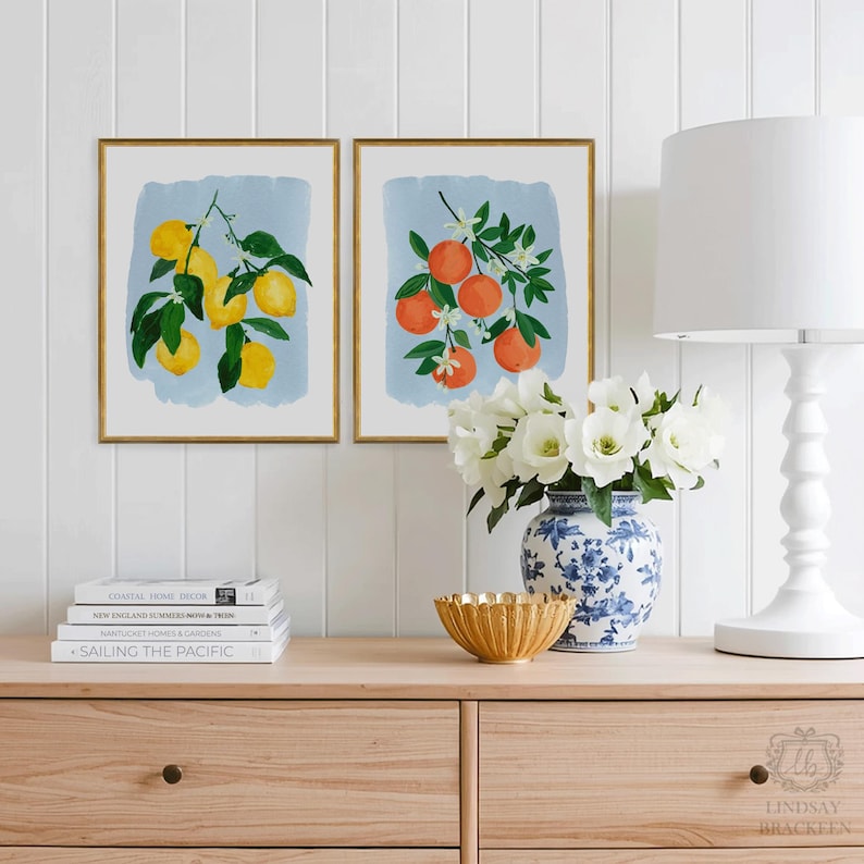 K&ouml;nnte beinhalten: Zwei gerahmte botanische Drucke mit Zitronen und Orangen auf hellblauem Hintergrund. Die Drucke haben goldene Rahmen und werden &uuml;ber einer Holzkommode ausgestellt. Eine wei&szlig;e Lampe, eine Blumenvase und B&uuml;cher sind ebenfalls vorhanden.