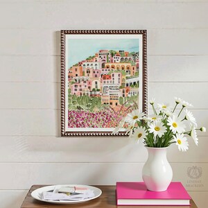 May include: A framed watercolor print depicts a colorful hillside town with pink, peach, and white buildings. The print is in a dark brown frame. A white vase with daisies sits on a wooden table, next to a stack of pink books.