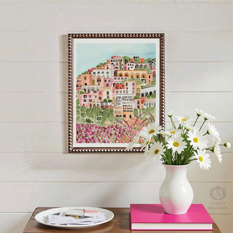 May include: A framed watercolor print depicts a colorful hillside town with pink, peach, and white buildings. The print is in a dark brown frame. A white vase with daisies sits on a wooden table, next to a stack of pink books.