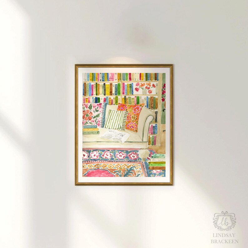 May include: Watercolour print depicting a comfortable reading space, featuring a bookshelf with vibrant books. A white sofa with patterned cushions rests on a floral rug. The artwork is framed in a gold-coloured frame.