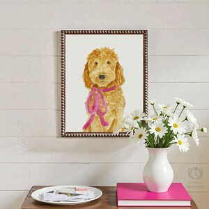 May include: A framed watercolor painting of a golden-colored dog with curly fur, wearing a bright pink ribbon tied around its neck. The artwork is displayed in a dark, textured frame and features a light-colored background behind the dog.