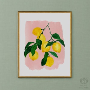 Op de afbeelding: Een ingelijste kunstdruk met een aquarelillustratie van een citroentak met felgele citroenen en donkergroene bladeren tegen een zachtroze penseelstreekachtergrond. Het kunstwerk is gevat in een dunne goudkleurige metalen lijst.