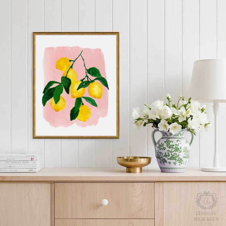 Op de afbeelding: Een ingelijste kunstdruk met een geschilderde illustratie van gele citroenen met groene bladeren op een zachtroze aquarelachtergrond. Het kunstwerk is ingelijst in een dunne gouden lijst en toont een botanisch thema met citrusvruchten.