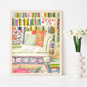 May include: Watercolour print depicting a reading nook with a bookshelf full of colourful books. A cream sofa with patterned cushions, an open book, and a floral rug are featured. The artwork is framed in a light wood frame.