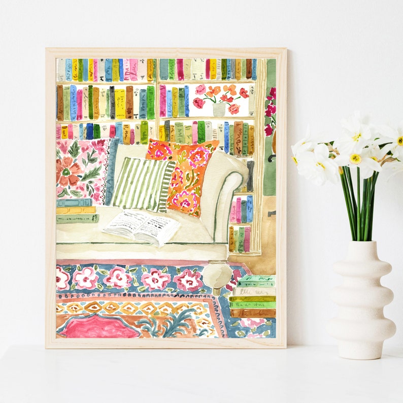 May include: Watercolour print depicting a reading nook with a bookshelf full of colourful books. A cream sofa with patterned cushions, an open book, and a floral rug are featured. The artwork is framed in a light wood frame.