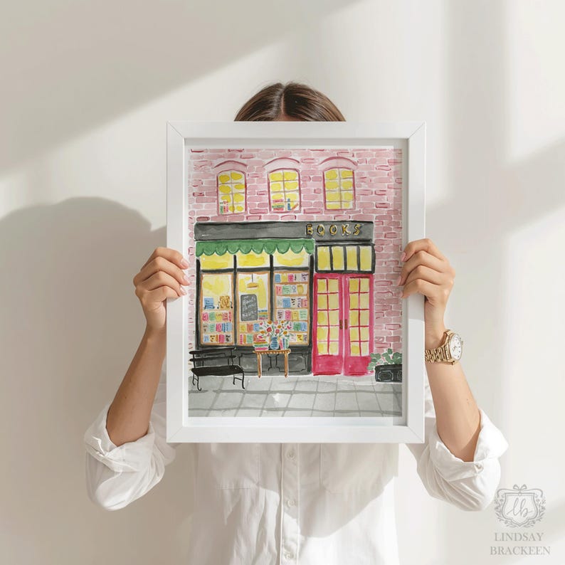 May include: Watercolor painting of a bookstore with a green awning and the word "BOOKS" above the entrance. The building has a red brick facade and three yellow-lit windows. A person holds the framed artwork.