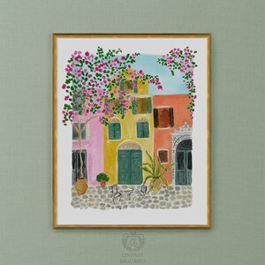 May include: A watercolor art print in a gold frame depicting a Mediterranean street scene with pink bougainvillea, a yellow building with green doors, an orange building, a cobblestone ground, two chairs, a small table, and a potted plant.