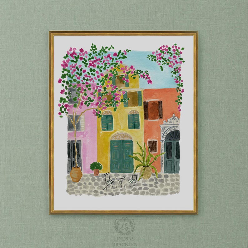 May include: A watercolor art print in a gold frame depicting a Mediterranean street scene with pink bougainvillea, a yellow building with green doors, an orange building, a cobblestone ground, two chairs, a small table, and a potted plant.