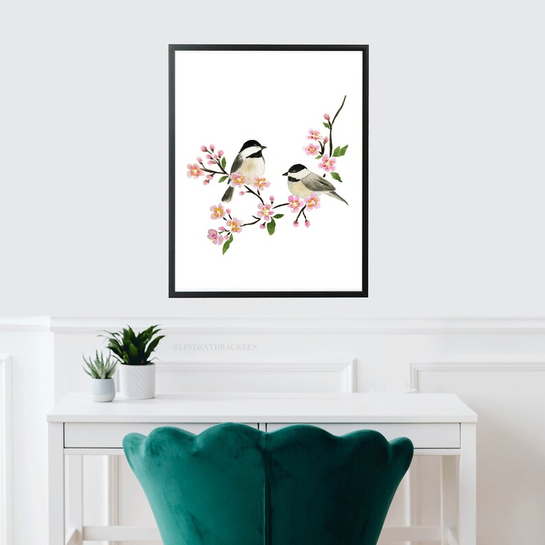 May include: A framed art print showing two small birds on a flowering branch with pink blossoms. The birds have black and white markings. The artwork is set against a white background and framed in a black border. The print is displayed above a white desk.