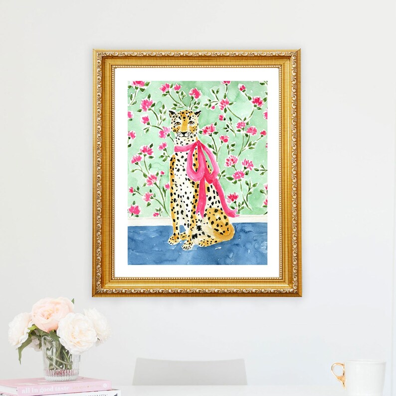May include: A framed watercolor painting of a cheetah wearing a pink ribbon. The cheetah is sitting on a blue background with a green and pink floral pattern behind it.