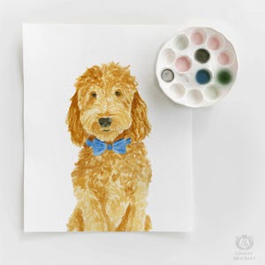 May include: A watercolor painting of a golden-brown dog with curly fur wearing a bright blue bow tie. Next to the painting is a white circular paint palette containing various colors of dried watercolor paint in small circular wells.