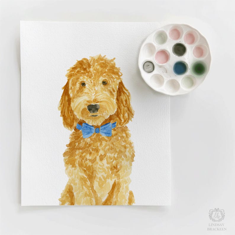 May include: A watercolor painting of a golden-brown dog with curly fur wearing a bright blue bow tie. Next to the painting is a white circular paint palette containing various colors of dried watercolor paint in small circular wells.